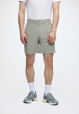 Dospelá osoba oblečená v svetlosivých kraťasoch s bočnými vreckami, bielych členkových ponožkách, sivých teniskách a bielom tričku s krátkym rukávom stojaca na jednoduchom pozadí.