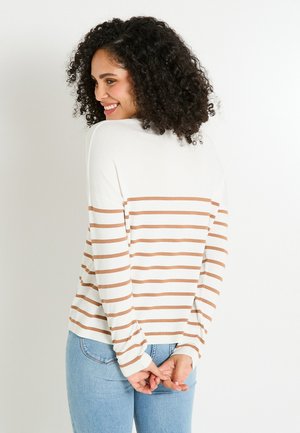 Femme aux cheveux bouclés et foncés souriant, portant un pull rayé blanc et marron et un jean bleu clair, se tenant avec les mains croisées derrière le dos.