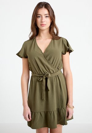 Frau trägt ein olivgrünes Wickelkleid mit kurzen Ärmeln, gebundenem Taillengürtel und gerafftem Saum, steht vor einem schlichten weißen Hintergrund.