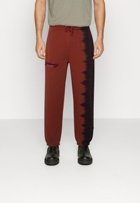 Burgunderfarbene Sweatpants mit einem enganliegenden Bündchen, die auf einer Seite ein schwarzes Batikmuster und auf der Vorderseite ein lila Logo aufweisen. Mit Kordelzugbund.