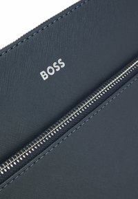 Granatowa skórzana saszetka o teksturowanej powierzchni, srebrzysty zamek błyskawiczny oraz wytłoczony na froncie napis "BOSS". Szwy na krawędziach zwiększają trwałość.
