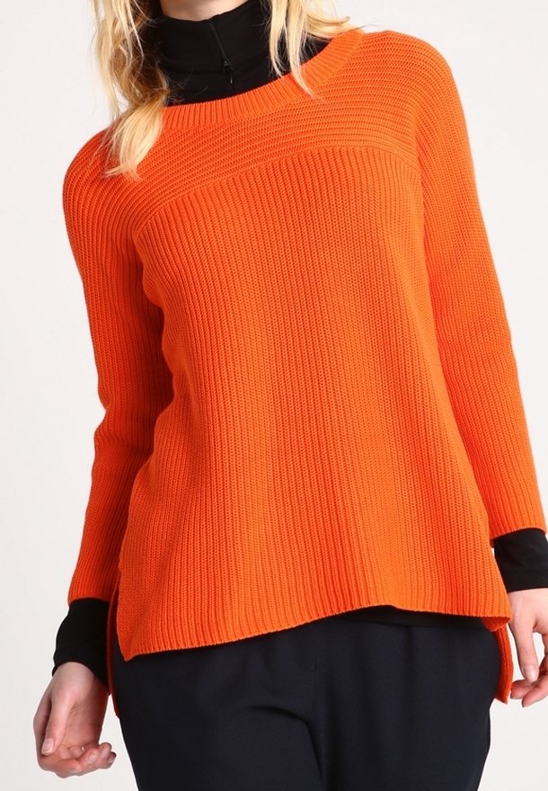 Femme portant un pull en maille côtelée orange vif superposé à un haut à manches longues noir avec un pantalon noir.