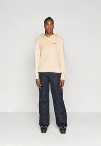 Une femme se tient face à l'avant, portant un sweat à capuche crème, un pantalon cargo noir et des bottes de ski noires, devant un fond clair uni.