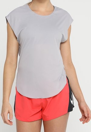 Frau trägt hellgraues ärmelloses Oberteil und rote Sportshorts mit schwarzen Seitenpaneelen, steht mit entspannten Armen seitlich am Körper.