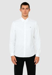 Camicia bianca a maniche lunghe realizzata in tessuto liscio con colletto classico, caratterizzata da un piccolo logo nero "AX" sul lato sinistro del petto.