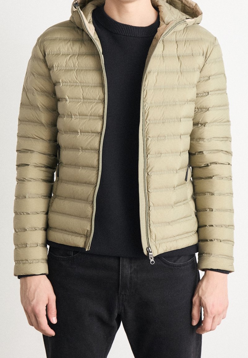 Knielange, khakifarbene Daunenjacke mit horizontalen Nähten, Kapuze und Reißverschluss. Über einem dunklen, strukturierten Pullover getragen.