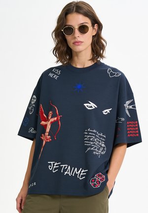 Junge Frau trägt runde Sonnenbrille und ein dunkelblaues oversized Hemd mit roten und weißen romantischen Drucken und Text.