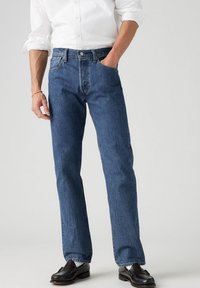 Vaqueros de mezclilla en azul medio, corte recto con cinco bolsillos y cierre de botones. Combinados con una camisa blanca y mocasines negros.