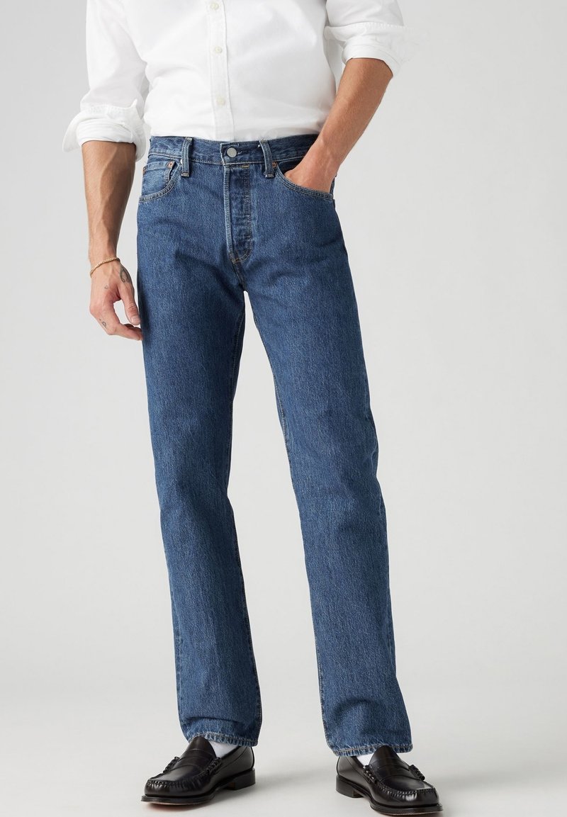 Vaqueros de mezclilla en azul medio, corte recto con cinco bolsillos y cierre de botones. Combinados con una camisa blanca y mocasines negros.