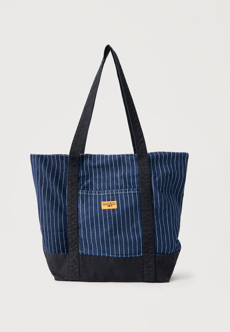 Τσάντα tote από ύφασμα με μπλε και μαύρες ρίγες, μαύρες μακριές λαβές και εξωτερική τσέπη με μικρή κίτρινη ετικέτα.