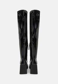 Bottes noires hauteur genou en cuir verni brillant. Elles présentent un talon carré et des fermetures éclair à l'arrière avec une texture lisse.