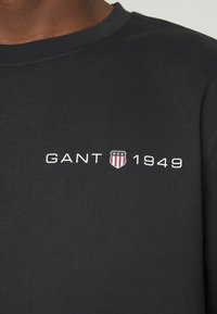 T-shirt nera a maniche lunghe con collo a girocollo, realizzata in cotone, con testo bianco "GANT 1949" e un piccolo logo a scudo in rosso e blu.