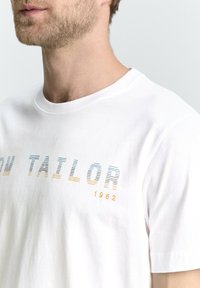 Camiseta de algodón blanca con un estampado de degradado horizontal en azul y naranja, que presenta el texto "TAYLOR" y "1962" en diferentes grosores de línea.
