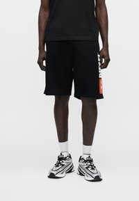 Άνδρας με μαύρο σορτς που έχει λευκό και πορτοκαλί λογότυπο Diesel στο αριστερό πόδι, λευκές κάλτσες και λευκά-μαύρα αθλητικά παπούτσια Diesel, στέκεται σε λευκό δάπεδο.