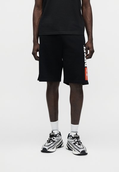 Homme portant un short noir avec le logo Diesel blanc et orange sur la jambe gauche, des chaussettes blanches et des baskets Diesel blanches et noires, debout sur un sol blanc.
