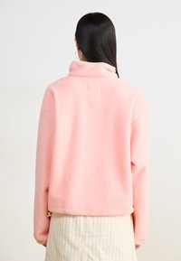 Pull en polaire rose avec un col montant, surface texturée et un petit patch carré dans le dos. En dessous, un pantalon rayé de couleur claire.