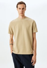 Beige T-Shirt mit kurzen Ärmeln und strukturierter Oberfläche, Rundhalsausschnitt, mit dezenten Nähdetails. Getragen mit dunklen Hosen.