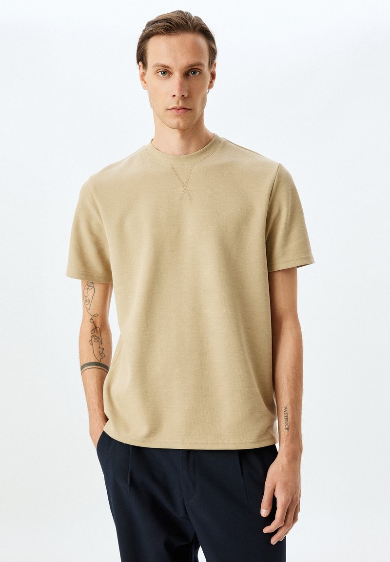 Beige T-Shirt mit kurzen Ärmeln und strukturierter Oberfläche, Rundhalsausschnitt, mit dezenten Nähdetails. Getragen mit dunklen Hosen.