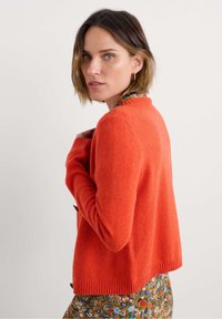 Gilet en laine rouge avec un ourlet et des poignets côtelés, présentant un col rond et des accents de boutons. Porté sur une jupe à motifs floraux.