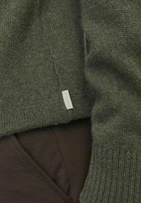 Suéter de punto verde oliva con puños acanalados, con una etiqueta de tela en la parte inferior. Se combina con pantalones oscuros de textura marrón.