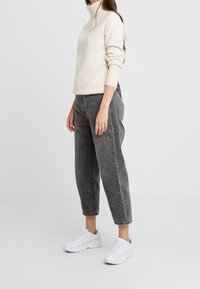 Kobieta nosząca jasnobeżowy sweter z golfem, wyblakłe szare spodnie o prostym kroju z wysokim stanem i kieszeniami oraz białe sportowe buty, stojąca.