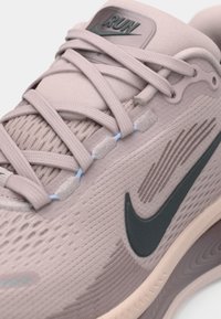 Jasnoróżowy but sportowy z teksturowaną siateczkową cholewką, szarym znakiem Nike, niebieskimi oczkami i płaskimi sznurówkami. Zbliżenie na szczegóły designu.