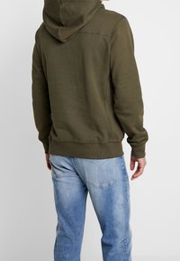 Olijfgroene capuchontrui met een zachte textuur en geribde zoom, gecombineerd met lichtblauwe spijkerbroek met achterzakken en een verweerde afwerking.