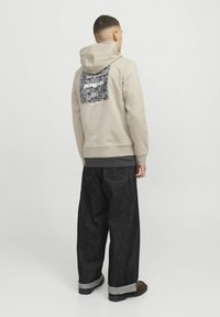 Beige hoodie with a patterned black patch on the back. Paired with loose-fitting dark denim jeans, rolled cuffs, and dark shoes.