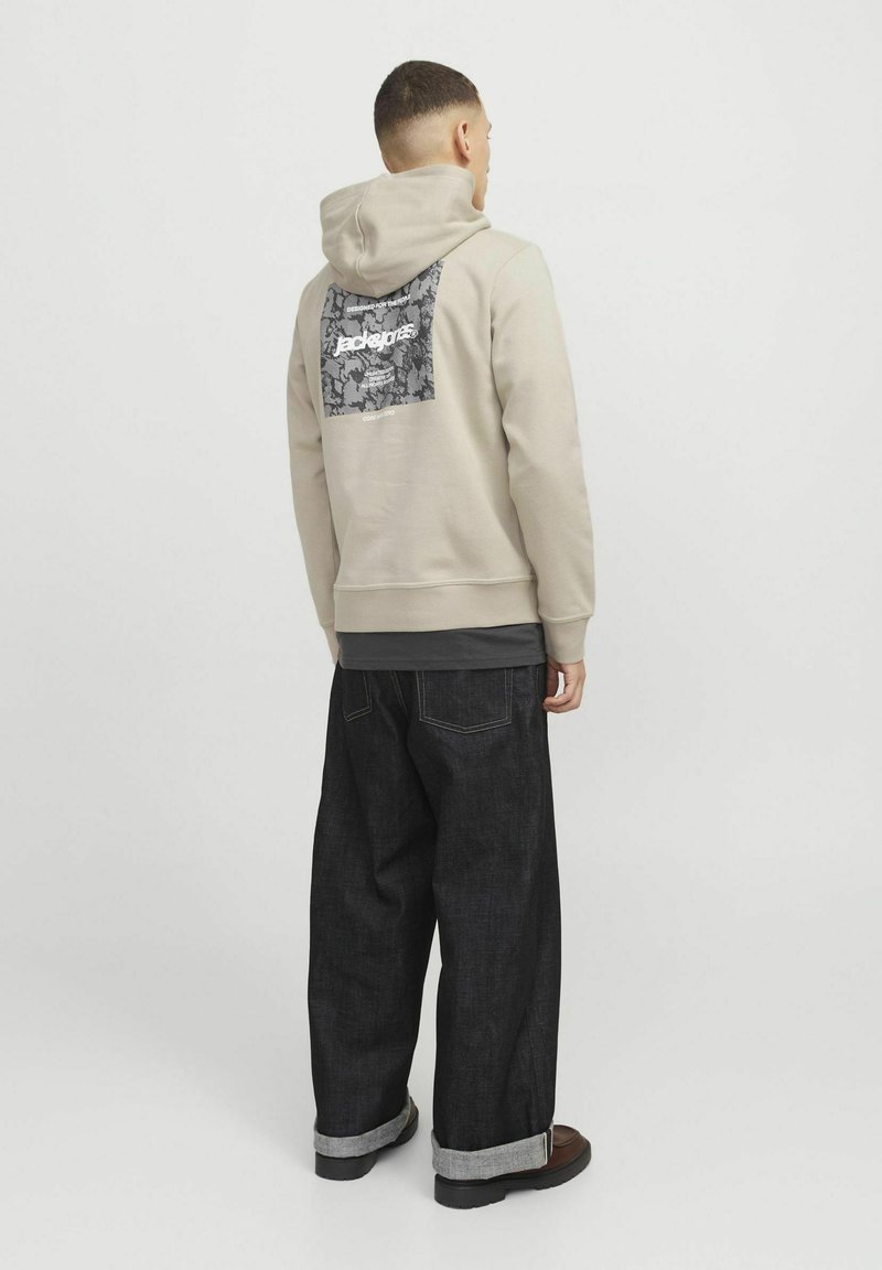Beige hoodie with a patterned black patch on the back. Paired with loose-fitting dark denim jeans, rolled cuffs, and dark shoes.