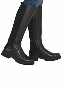 Altas botas de cuero negro con cremallera lateral y paneles de estiramiento acanalados, llevadas sobre jeans azules.