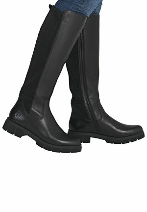 Bottes noires en cuir hautes avec fermeture éclair sur le côté et panneaux extensibles côtelés, portées sur un jean bleu.