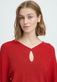 Crvena pletena džemper s V-izrezom koji ima mali otvor u obliku ključa. Dugi rukavi i teksturirani materijal. Jednostavan dizajn bez dodatnih ukrasa.