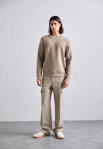 Young man with shoulder-length dark hair stands wearing a beige sweatshirt, khaki cargo pants, and cream sneakers on a gray floor.