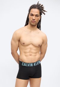 Μυώδης άνδρας με ντρέντλοκς που φοράει μαύρο εσώρουχο Calvin Klein στέκεται μπροστά σε απλό ανοιχτό φόντο, με τα χέρια πίσω από την πλάτη.