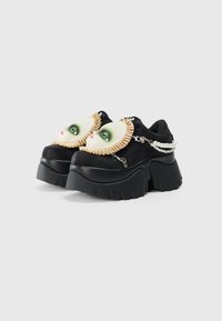 Sneakers platform neri con grandi decorazioni a forma di volto di bambola, rouches beige e accenti di catena di perle su uno sfondo bianco semplice.