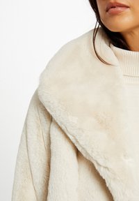 Kremowy sztuczny futro płaszcz z szerokim, zawijanym kołnierzem. Miękka tekstura i lekko nieregularny wzór na powierzchni. Warstwowy wygląd z przytulnym swetrem pod spód.