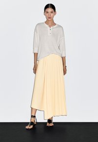 Jasnoszary top henley z rękawami do trzech czwartych, w połączeniu z plisowaną, sięgającą kostek, pastelowożółtą spódnicą. Czarne płaskie sandały.