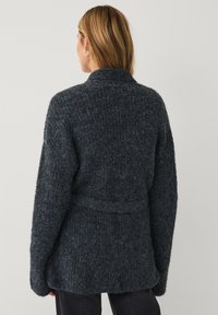 Frau mit blonden Haaren, die einen dunkelgrauen, flauschigen Strickcardigan mit Gürtel trägt, von hinten vor einem schlichten Hintergrund gezeigt.