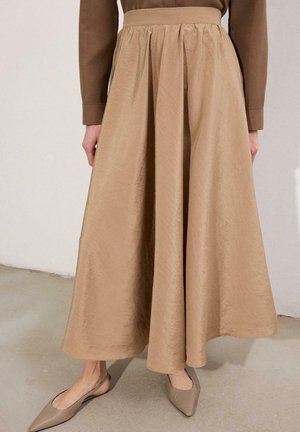 Jupe maxi beige à taille froncée associée à des ballerines slingback beige à bout pointu, posées sur un sol en béton clair.