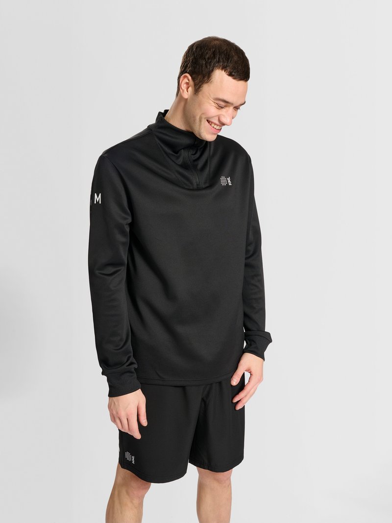 Jeune homme souriant en pull noir à manches longues et short, regardant vers le bas, debout devant un fond gris clair uni.