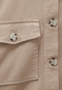 Tissu beige avec rabat de poche cousu et trois boutons beiges à motifs marbrés foncés sur un vêtement.