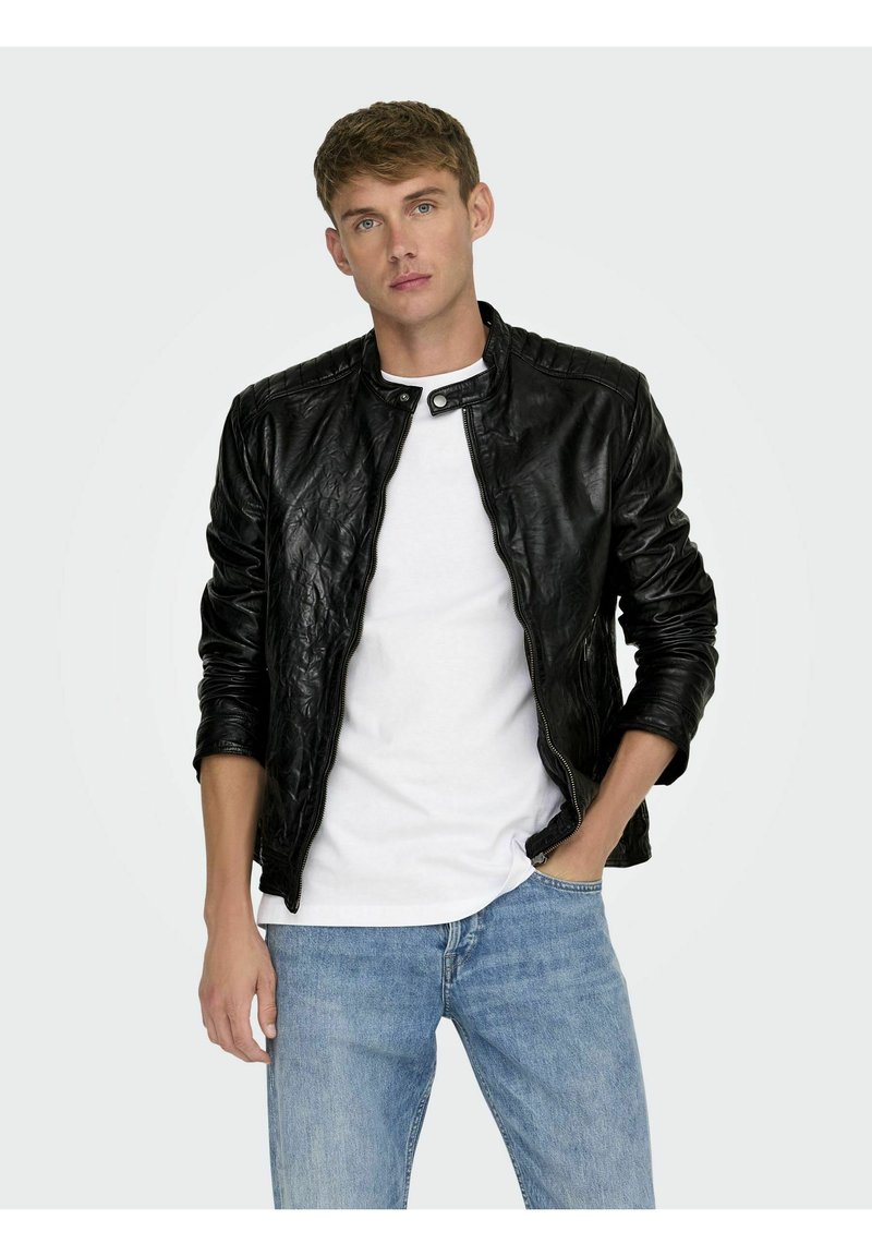 Zwarte leren jas met een ritssluiting aan de voorkant, ronde kraag en geribbelde schouderdetails, gedragen over een wit T-shirt en lichtblauwe jeans.