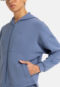 Svetlomodrá mikina na zips vyrobená z jemného materiálu. Má malé logo na hrudi, predné vrecká a voľný strih s kapucňou.