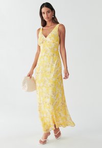 Yellow floral maxi dress with a deep V-neck, sleeveless design, flowing fabric, and a subtle sheen. Paired with a white handbag.