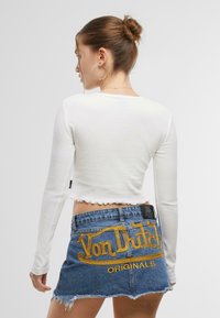 Vit långärmad crop top med en vågig fåll, kombinerad med en urblekt denim mini-kjol som pryds av gula broderade "Von Dutch Originals."