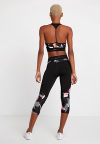 Donna con capelli biondi corti che indossa un reggiseno sportivo nero e leggings cropped a vita alta con motivi geometrici colorati, in piedi di spalle.