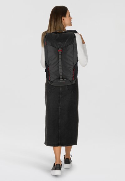 Mochila negra con material acolchado, acentos rojos y un asa superior. Tiene dos bolsillos laterales y correas ajustables. Usada por una persona vista desde atrás.