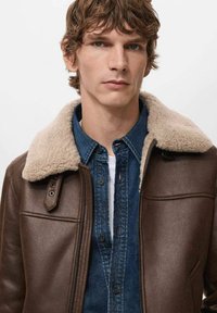Giacca di pelle marrone con colletto in shearling crema, chiusura a zip e tasca frontale, indossata sopra una camicia di jeans blu.