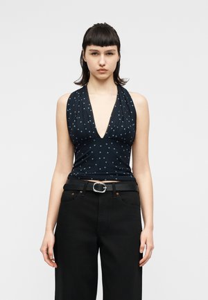 Γυναίκα με κοντά σκούρα μαλλιά που φοράει μπλε σκούρο μπλουζάκι χωρίς μανίκια με πουά και βαθύ V-λαιμόκοψη, καθώς και μαύρο παντελόνι ψηλόμεσο με ζώνη.