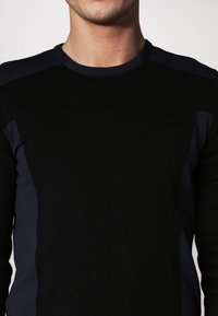 Pull noir en maille avec un col rond, présentant des panneaux latéraux bleu foncé. La texture est lisse et ajustée, avec une bordure côtelée au niveau du col.